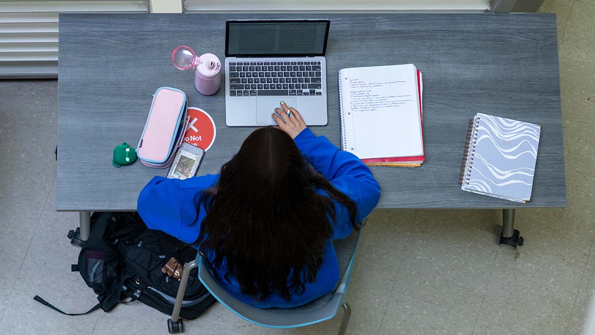 Overhead view of a student working at a desk, using a laptop with notebooks and a phone nearby.